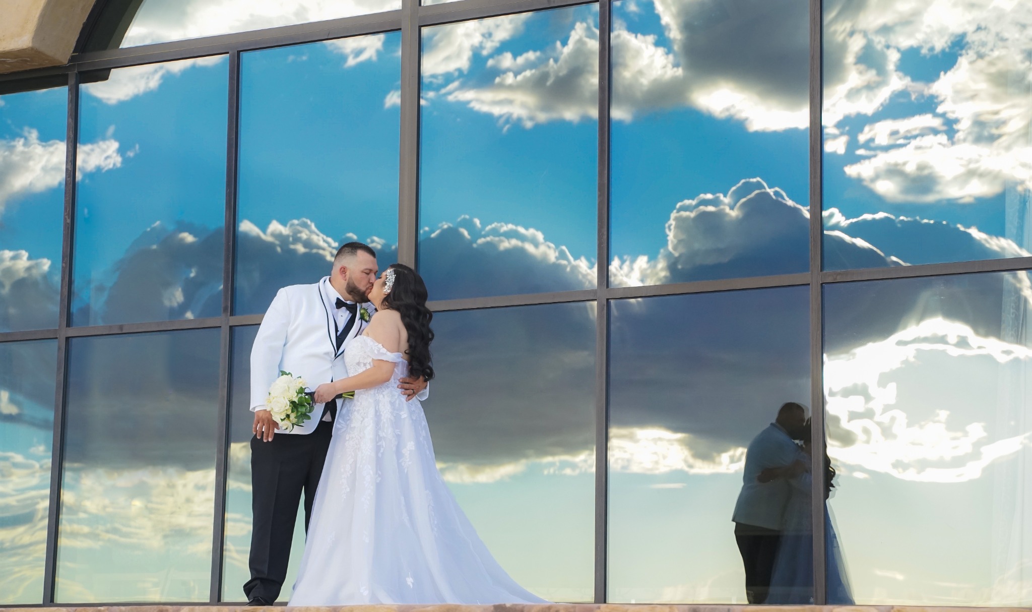 A bride and groom kissing beneath windows reflecting a blue sky and clouds